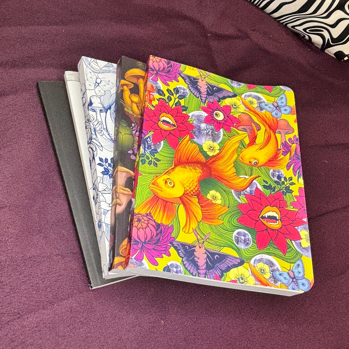 A stack of notebooks with colorful covers, featuring a prominent yellow and orange koi fish pattern on the top cover, placed on a dark purple fabric surface.