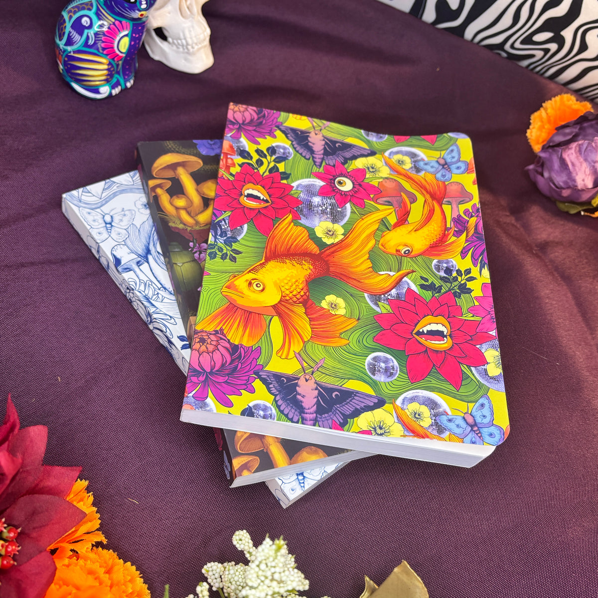 A stack of colorful illustrated journals lies on a dark purple fabric surface, surrounded by decorative flowers and small objects. The top notebook features a vivid design with orange and yellow koi fish, pink flowers, butterflies, and bubbles on a green background.