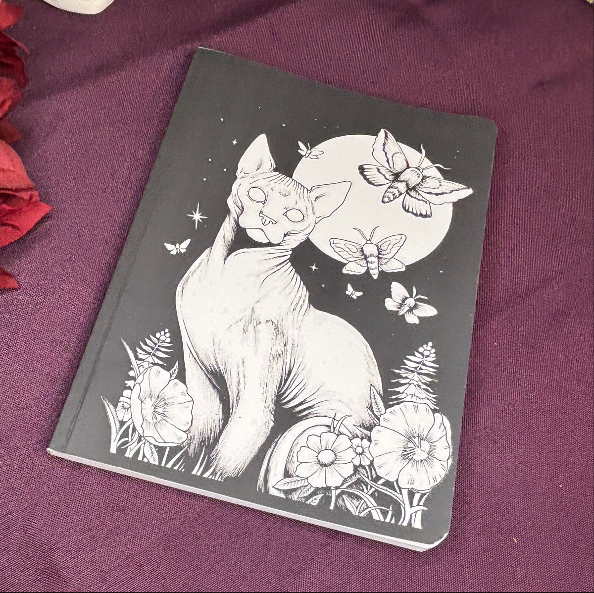 A black notebook or journal featuring a white line art illustration of a cat under a full moon, placed on a dark purple fabric surface with decorative props around it.