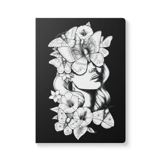 A front-facing view of a rectangular softcover journal with rounded corners, featuring a detailed black and white ink illustration against a solid black background. The design depicts a woman's face surrounded by large moths and blooming hibiscus flowers, with the moths covering her eyes to create a surreal and mystical look.