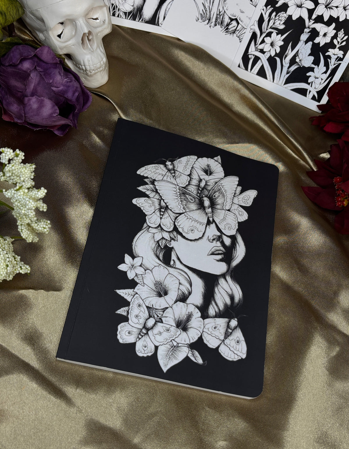 A black notebook with a white line-art illustration of a woman surrounded by flowers, styled on a gold satin fabric with a skull, purple flower, and black-and-white art prints.