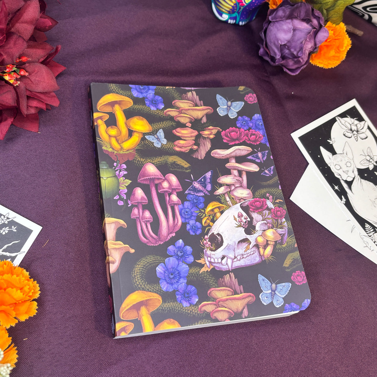 A notebook with a dark cover decorated with colorful mushrooms, flowers, butterflies, and a skull illustration, styled on a purple fabric surface with decorative flowers and printed cards.