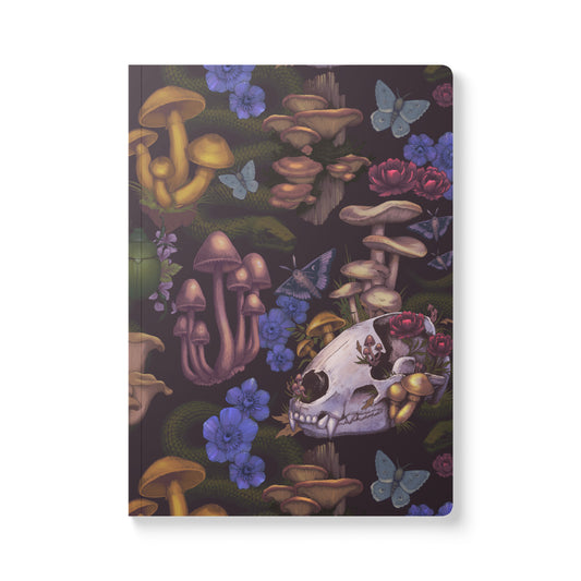 A front-facing view of a rectangular softcover journal with rounded corners, featuring a detailed, dark woodland-themed illustration pattern. The design includes realistic mushrooms, small white animal skulls, vibrant blue butterflies, and deep purple wildflowers spread across a solid dark grey background.