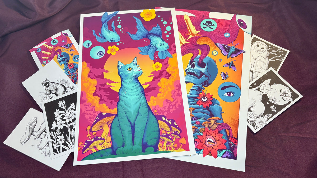 A set of colorful art prints and black-and-white sketch cards arranged on a dark purple fabric background. The main prints feature vibrant psychedelic illustrations, including a teal cat, blue fish, a blue skull, and floral motifs.