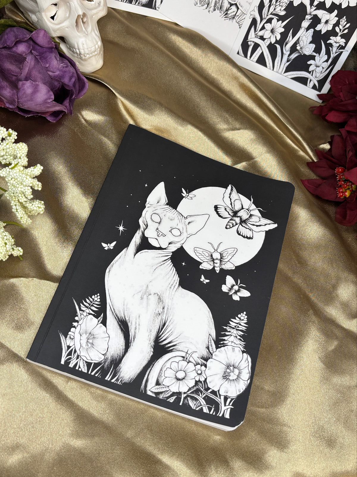 A black notebook with a white line art illustration of a cat under a full moon, styled on a gold satin fabric with a skull, flowers, and floral art sheets in the background.