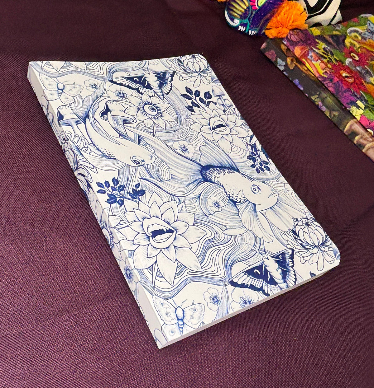 A notebook with a white cover decorated with blue line art of koi fish, lotus flowers, and butterflies, resting on a dark purple textured fabric surface.