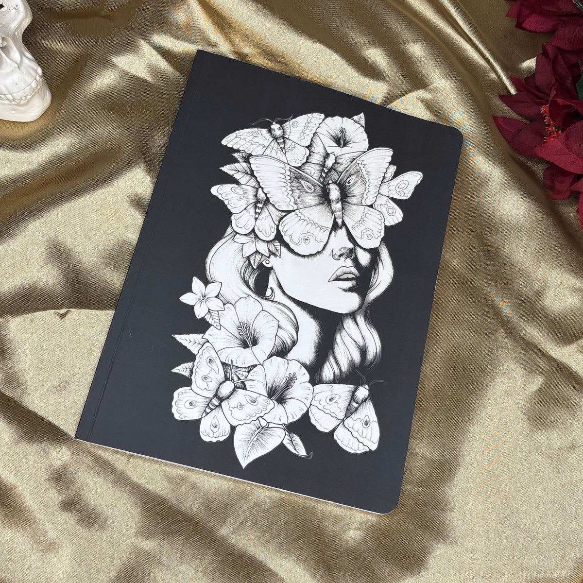 A black notebook with a detailed black and white illustration of a woman's face surrounded by flowers and butterflies, resting on a draped gold satin fabric with red flowers in the corner.