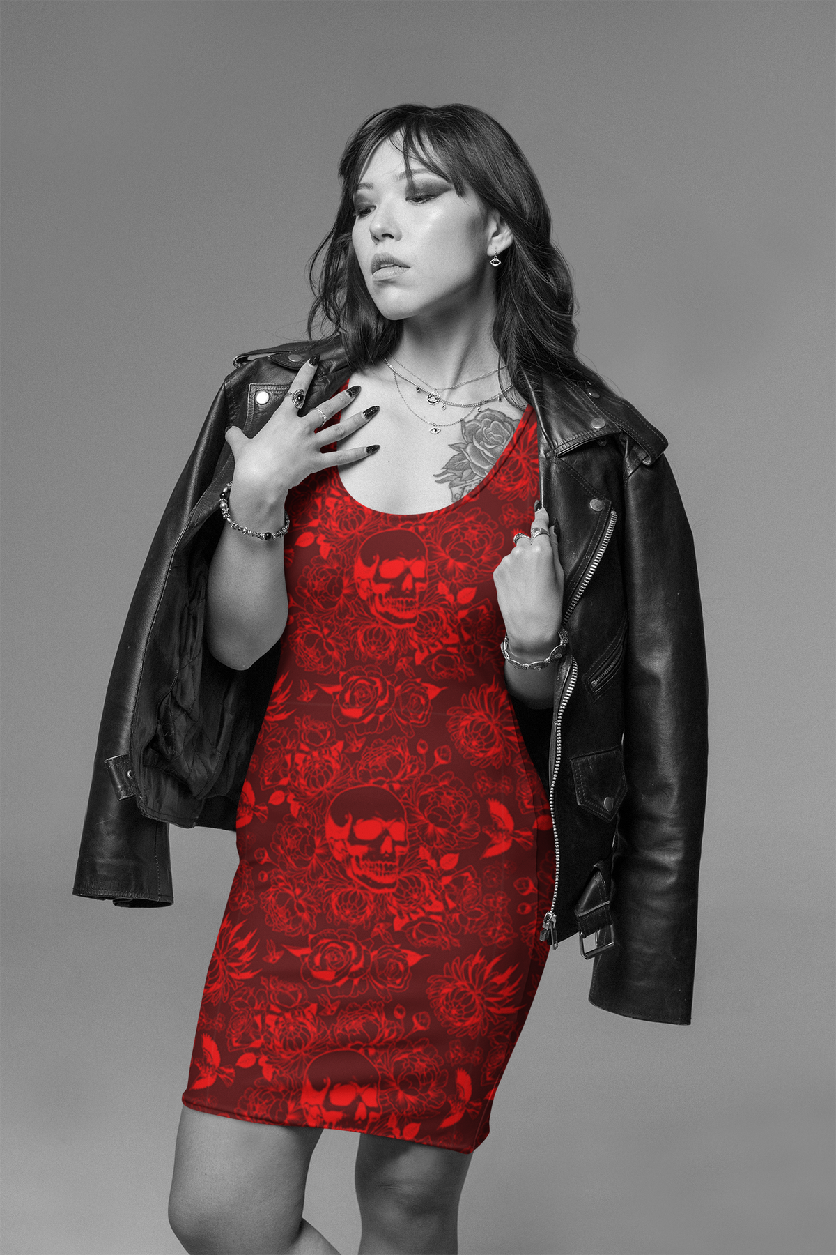 A woman posing in a black leather jacket over a red mini dress with a skull and rose pattern, set against a solid gray studio background.