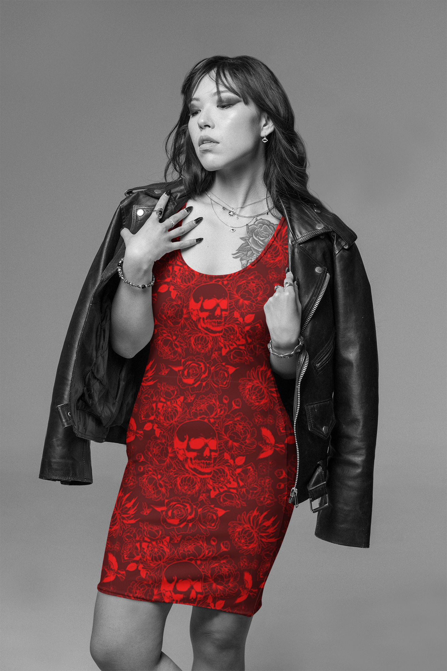 A woman posing in a black leather jacket over a red mini dress with a skull and rose pattern, set against a solid gray studio background.