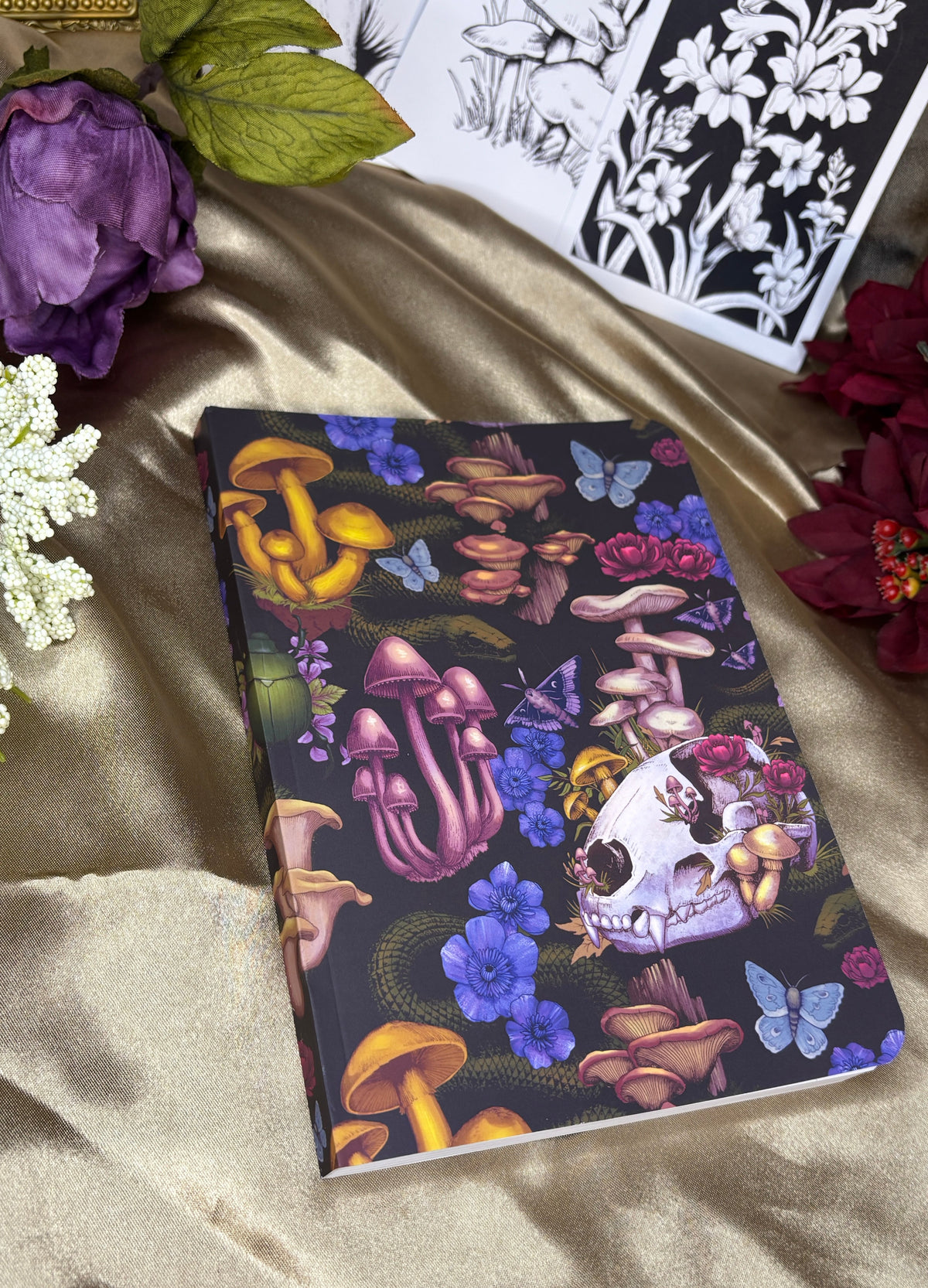 A softcover notebook with a dark cover decorated with colorful illustrations of mushrooms, skulls, flowers, and butterflies, styled on a gold satin fabric with decorative flowers.