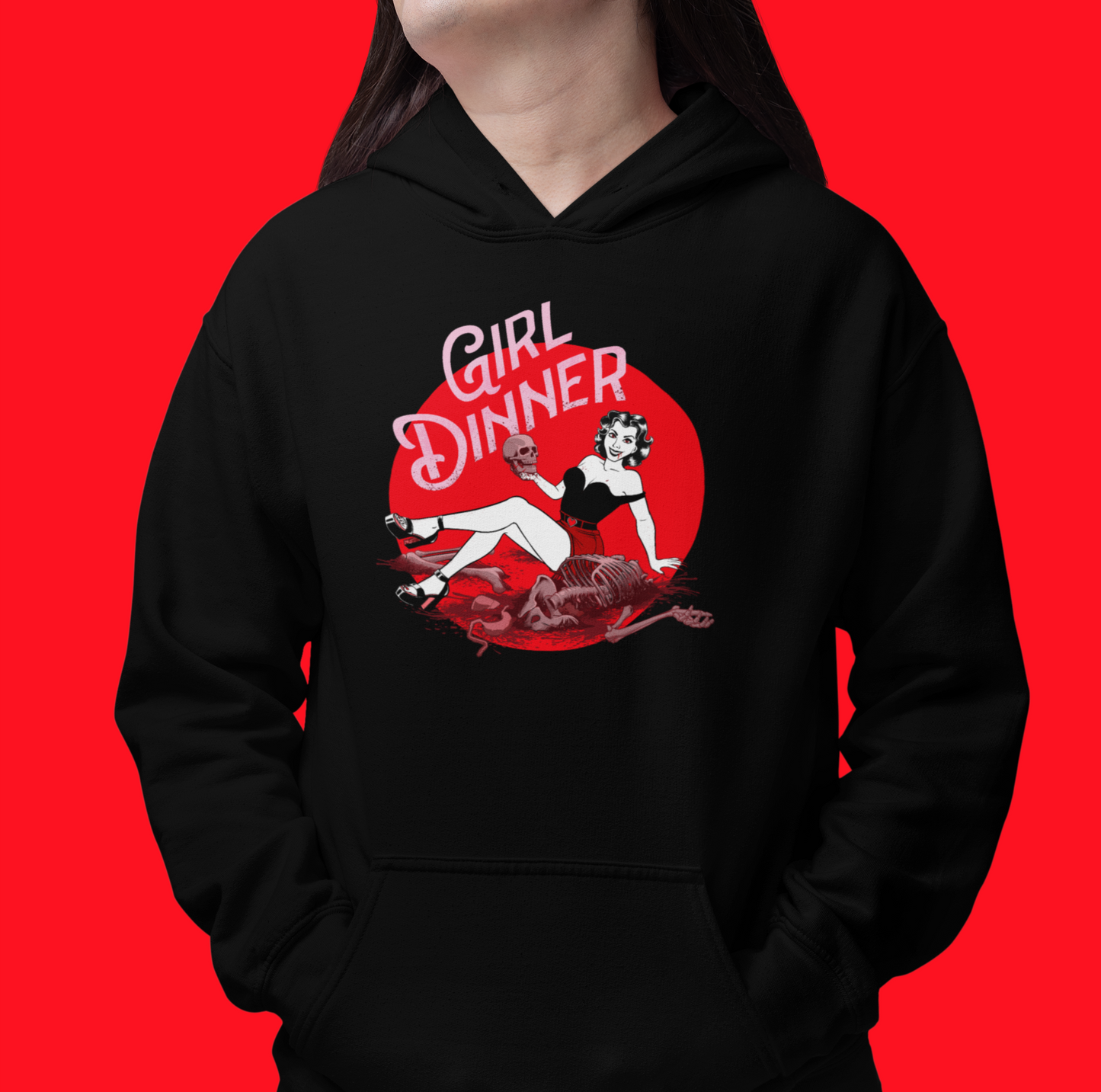 A person wearing a black pullover hoodie with a red and white retro-style graphic on the chest that reads 'GIRL DINNER' with an illustrated woman and a severed head, posed against a solid bright red background.