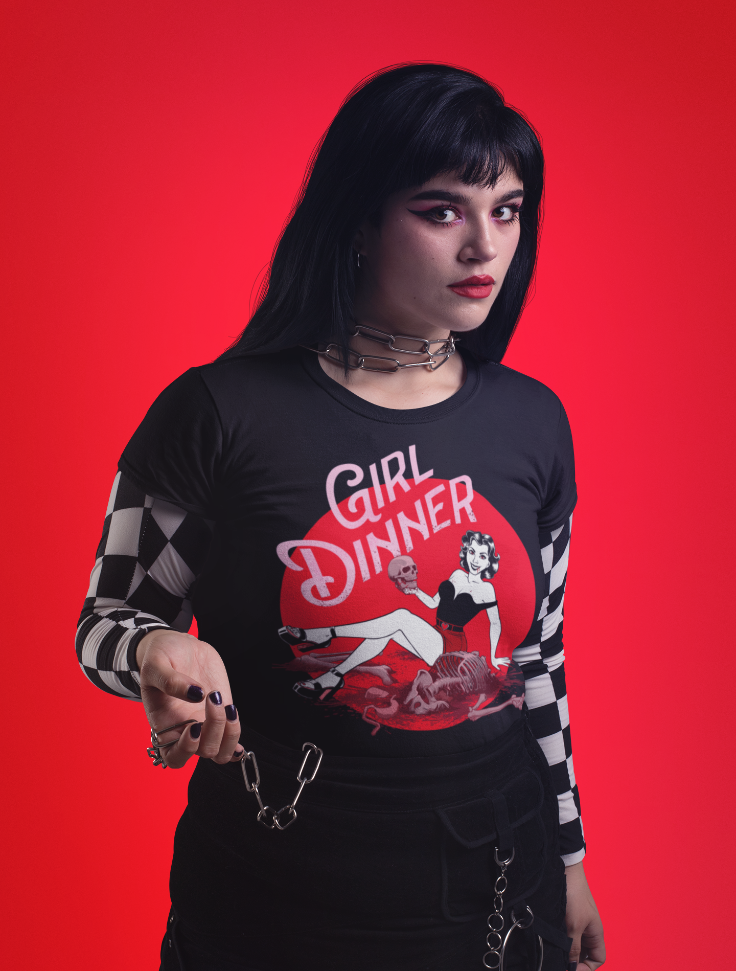 A model wearing a black long-sleeve graphic t-shirt with a red and white 'Girl Dinner' pin-up illustration, styled with black-and-white checkered sheer sleeves, a black skirt, and accessories against a solid red background.
