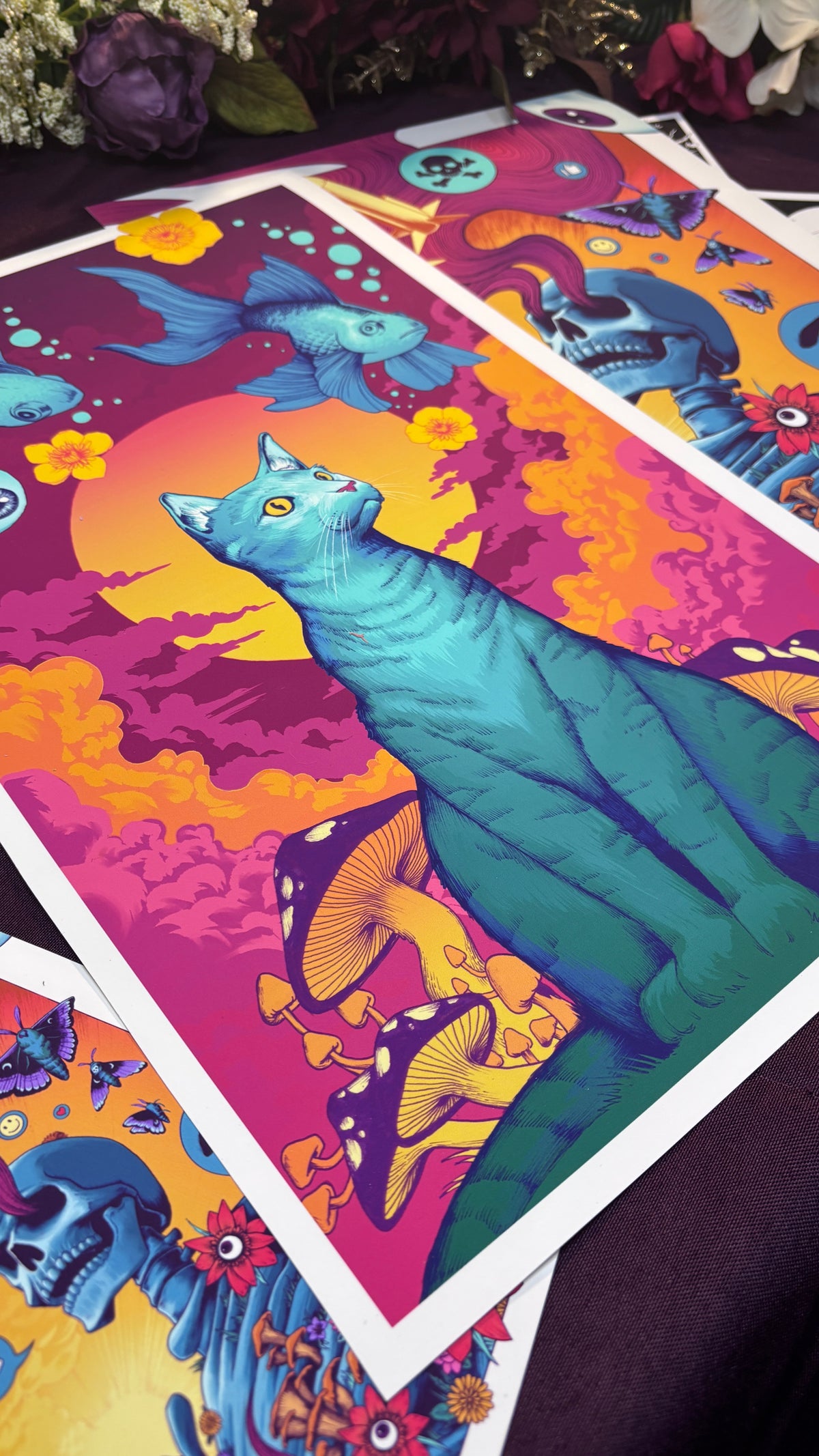 A colorful psychedelic art print featuring a large blue cat under a pink and orange sun, displayed on a dark purple surface with other prints partially visible.