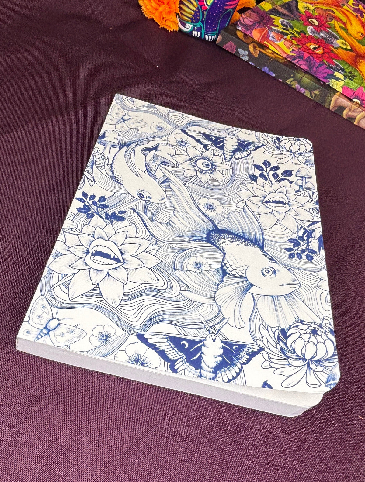 A notebook with a white cover decorated with blue line art of koi fish, lotus flowers, and butterflies, resting on a dark purple fabric surface.