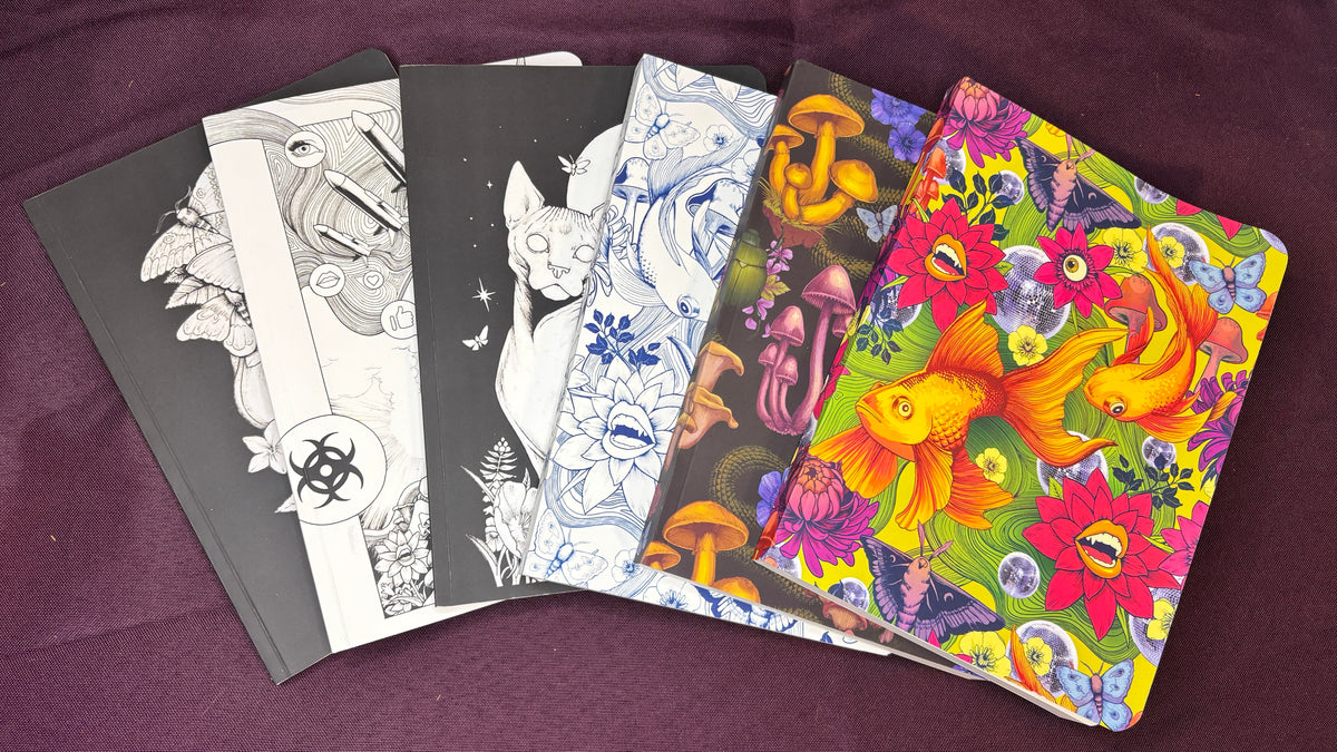 A fan arrangement of five illustrated notebooks or art books on a dark purple fabric background. The covers feature black-and-white line art, blue-and-white floral/animal sketches, orange-and-black mushroom artwork, and a vivid multicolor underwater-style design with goldfish, flowers, and sea life.