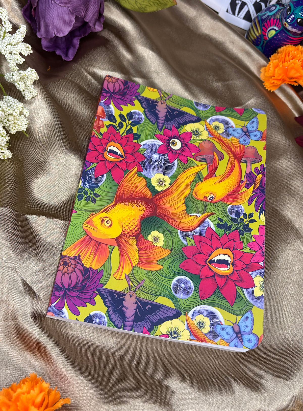 A notebook with a vibrant, colorful print of goldfish, lotus flowers, and butterflies, styled on a draped beige satin fabric with decorative flowers.
